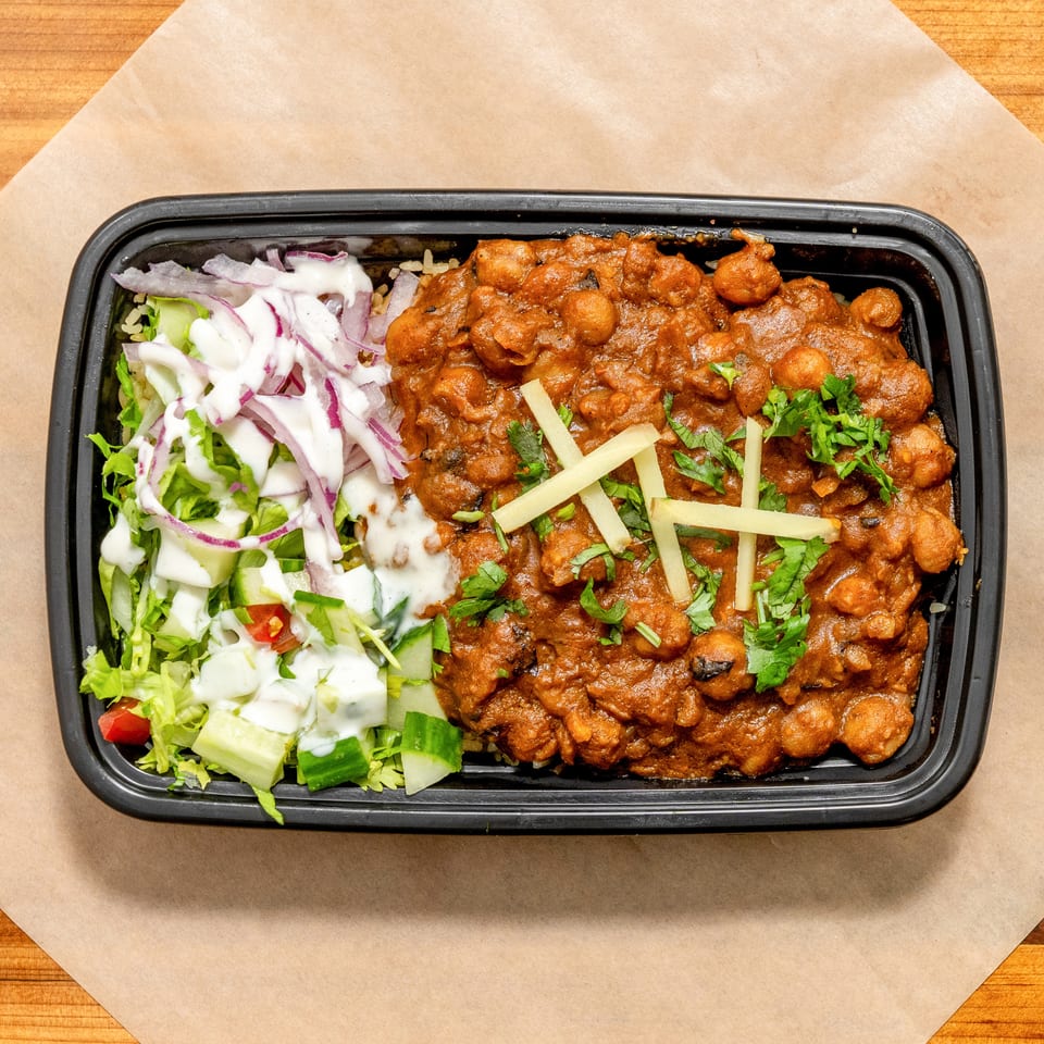 Takeout tray of creamy chickpea curry with rice, ginger, cilantro, and dressed salad on a wooden table.