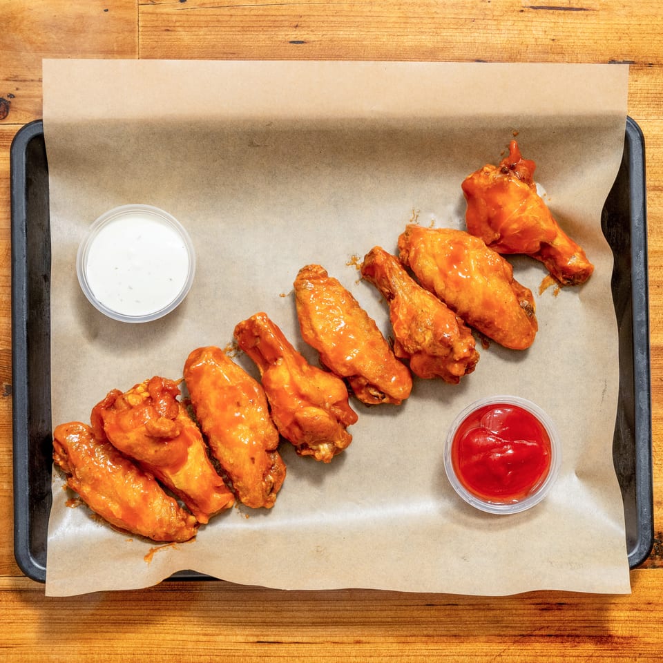 Buffalo chicken wings arranged on a parchment-lined tray with ranch and ketchup dipping sauces on a wooden table.