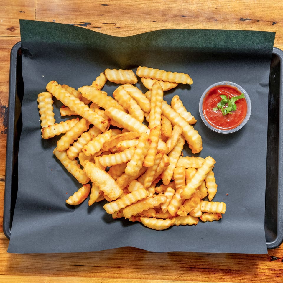 Crispy crinkle-cut fries piled on a black serving tray with a cup of tomato dipping sauce garnished with cilantro.