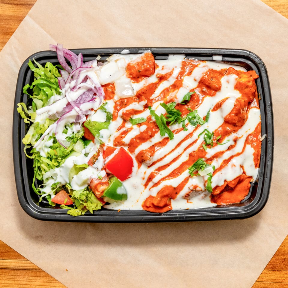 A black takeaway tray holds red-sauced grilled chicken drizzled with white sauce, alongside lettuce, onions, peppers, tomatoes, and herbs.