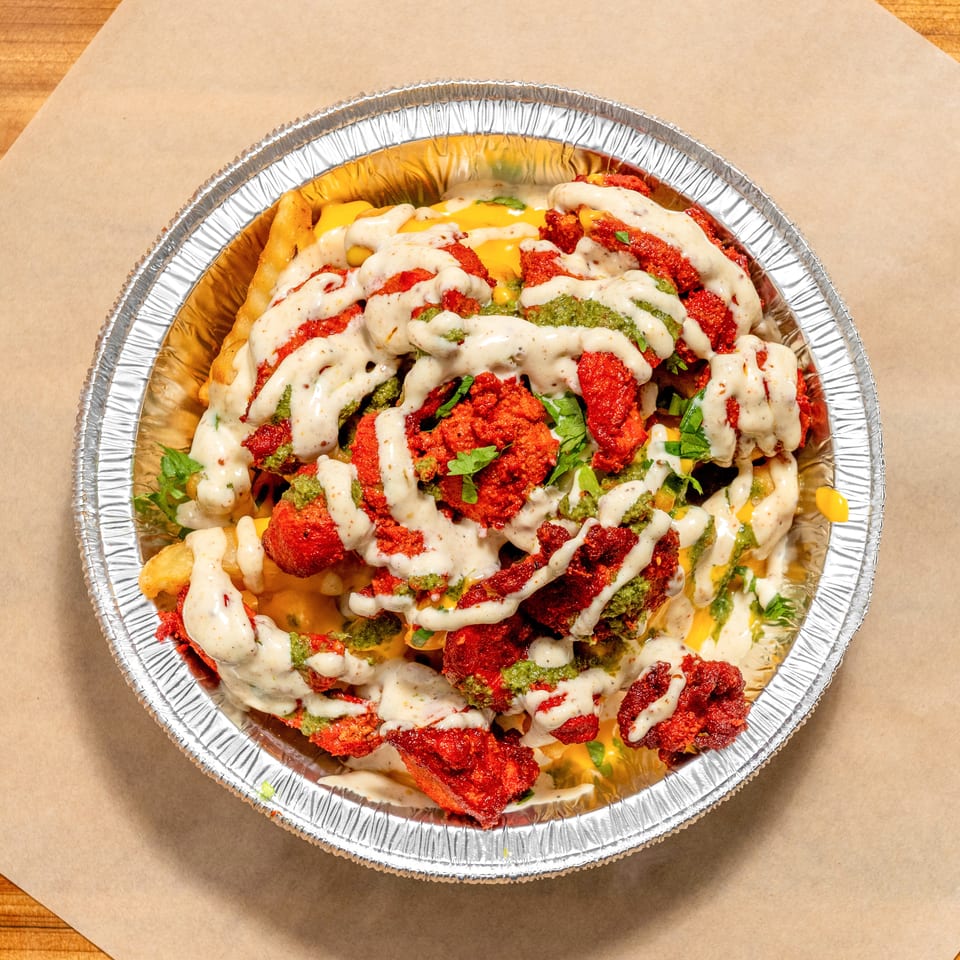 Loaded fries topped with crispy red chicken, melted cheese, green chutney, creamy sauce, and fresh herbs in a foil tray.