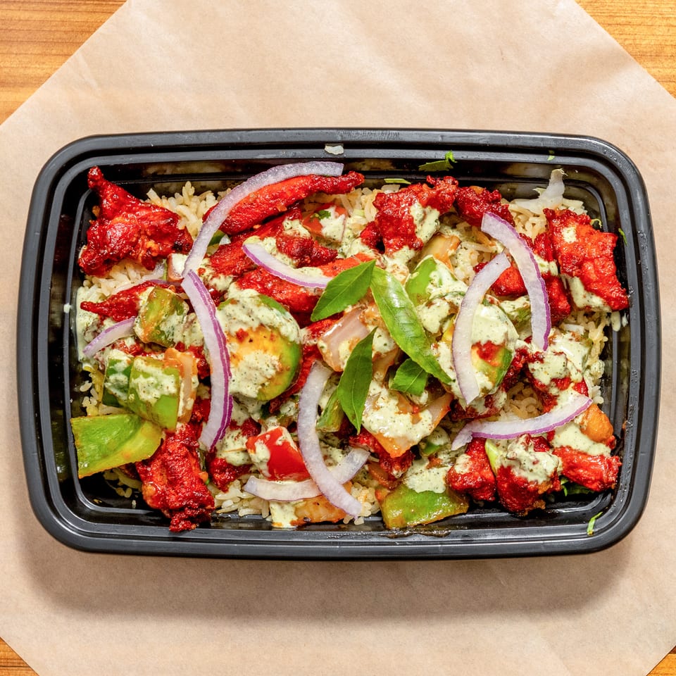 Black tray of spiced red chicken over rice, topped with avocado, peppers, red onion, herbs, and creamy green sauce.