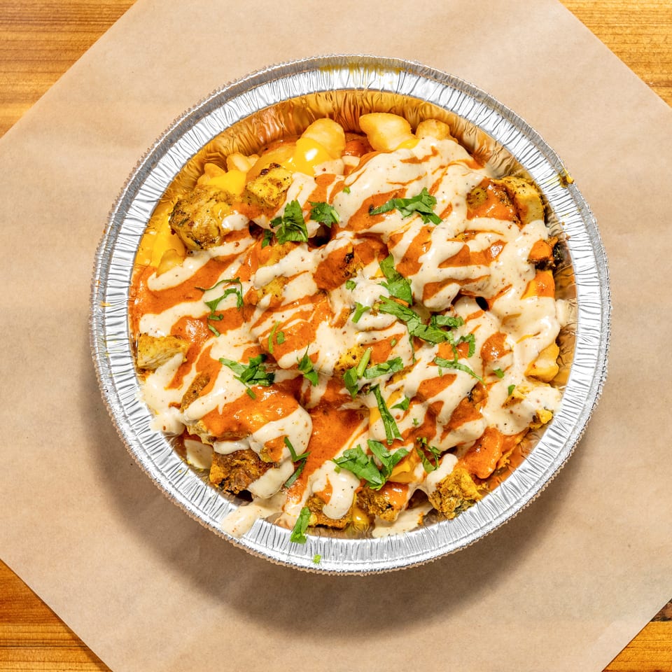 Loaded foil tray of seasoned grilled chicken and fries topped with orange and creamy sauces, melted cheese, and chopped herbs.